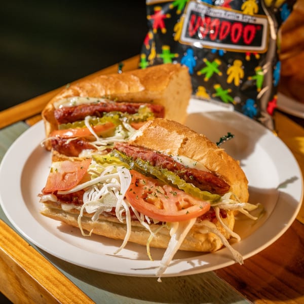 Two Nola Voodoo Tavern creole sausage po'boys with toppings and a bag of Voodoo chips.