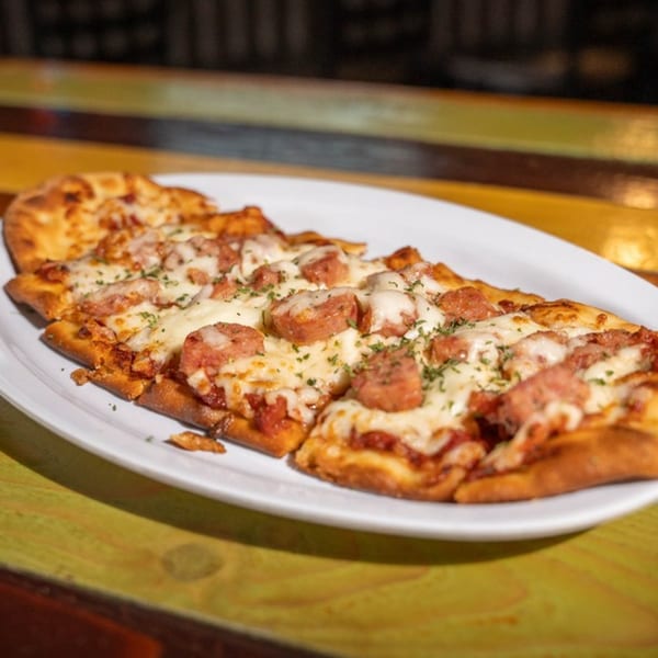 Nola Voodoo Tavern's flatbread pizza topped with melted cheese, sausage, and herbs on a white plate.